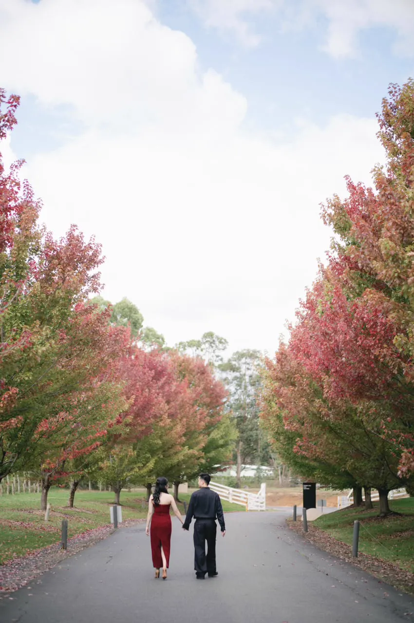 Vivi in a red dress and Max walking down a road lined with autumn trees