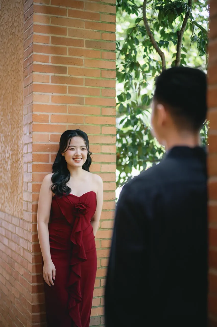 Vivi in a red dress smiling against a brick wall while Max looks on