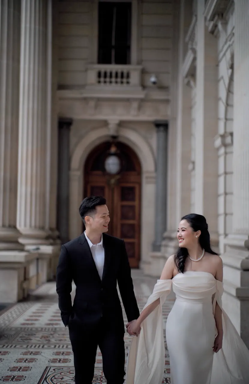 Vivi in a white gown and Max in a black suit walking through a stone colonnade, smiling at each other